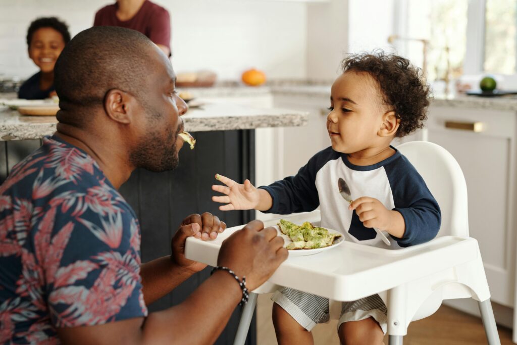 Father feeding toddler in high chair at home with family in kitchen.